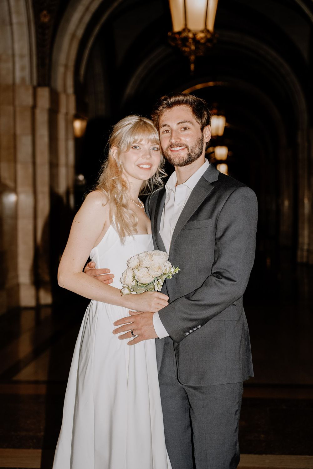 A bride in a white dress and groom in a gray suit embrace in an elegant arched hallway with vintage lighting.