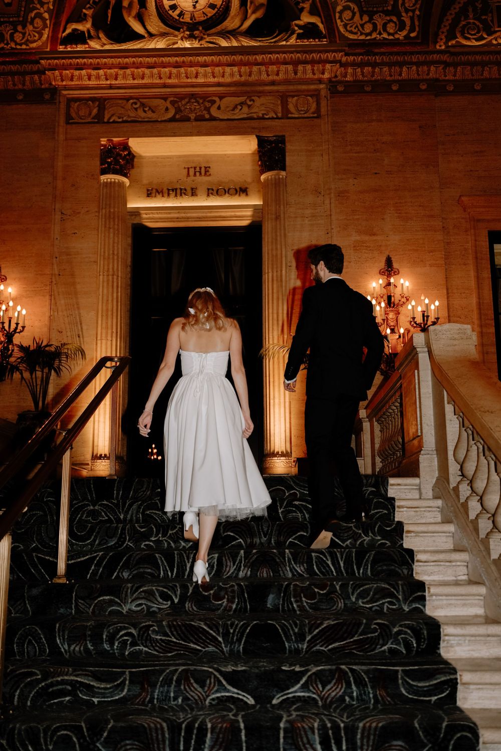 Wedding ceremony setup in an elegant ballroom with ornate columns and rows of black chairs illuminated by candlelight.
