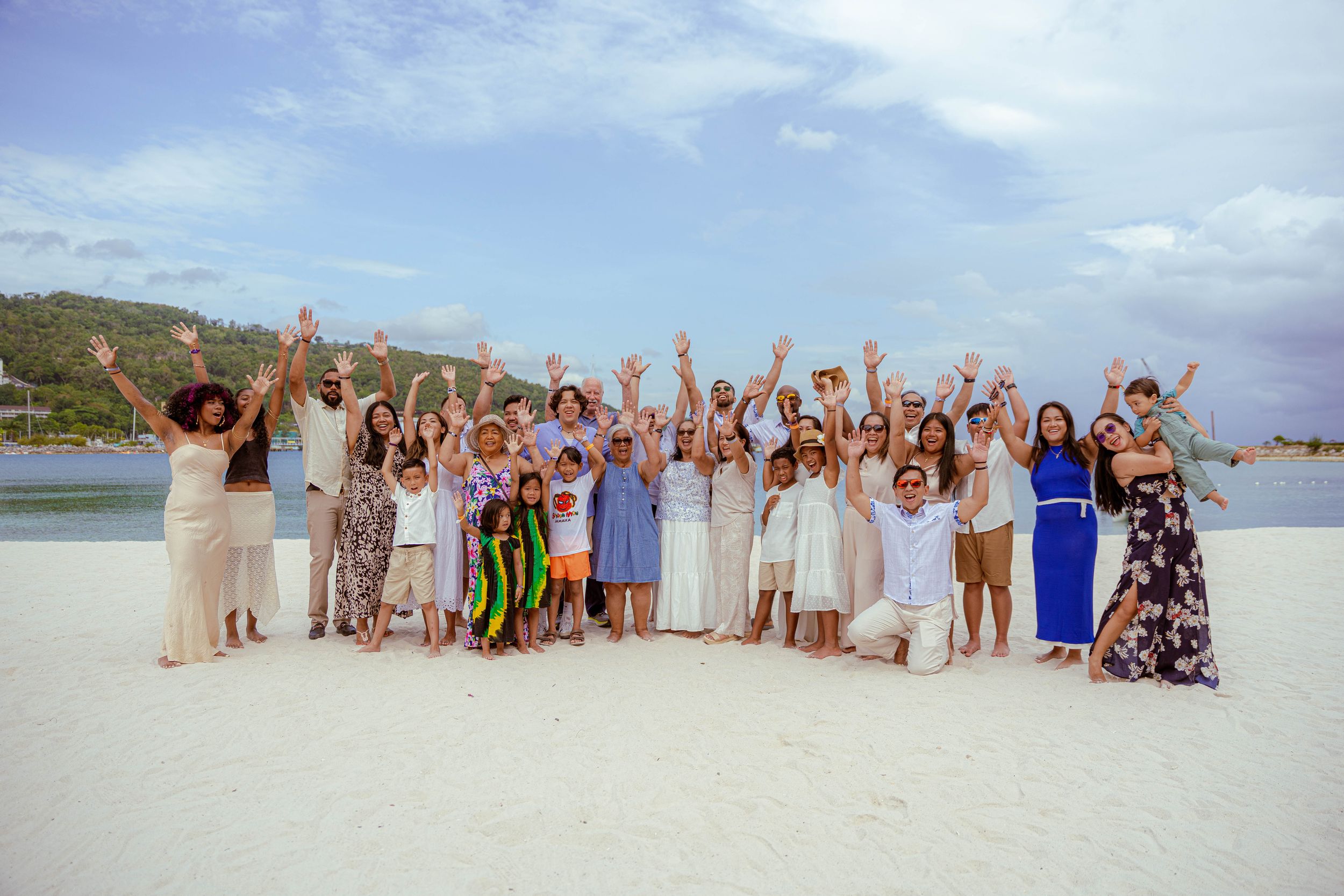 Jamaican Family Photograph large group of celebratory people with raised arms stand together on a white sand beach with blue water and sky.