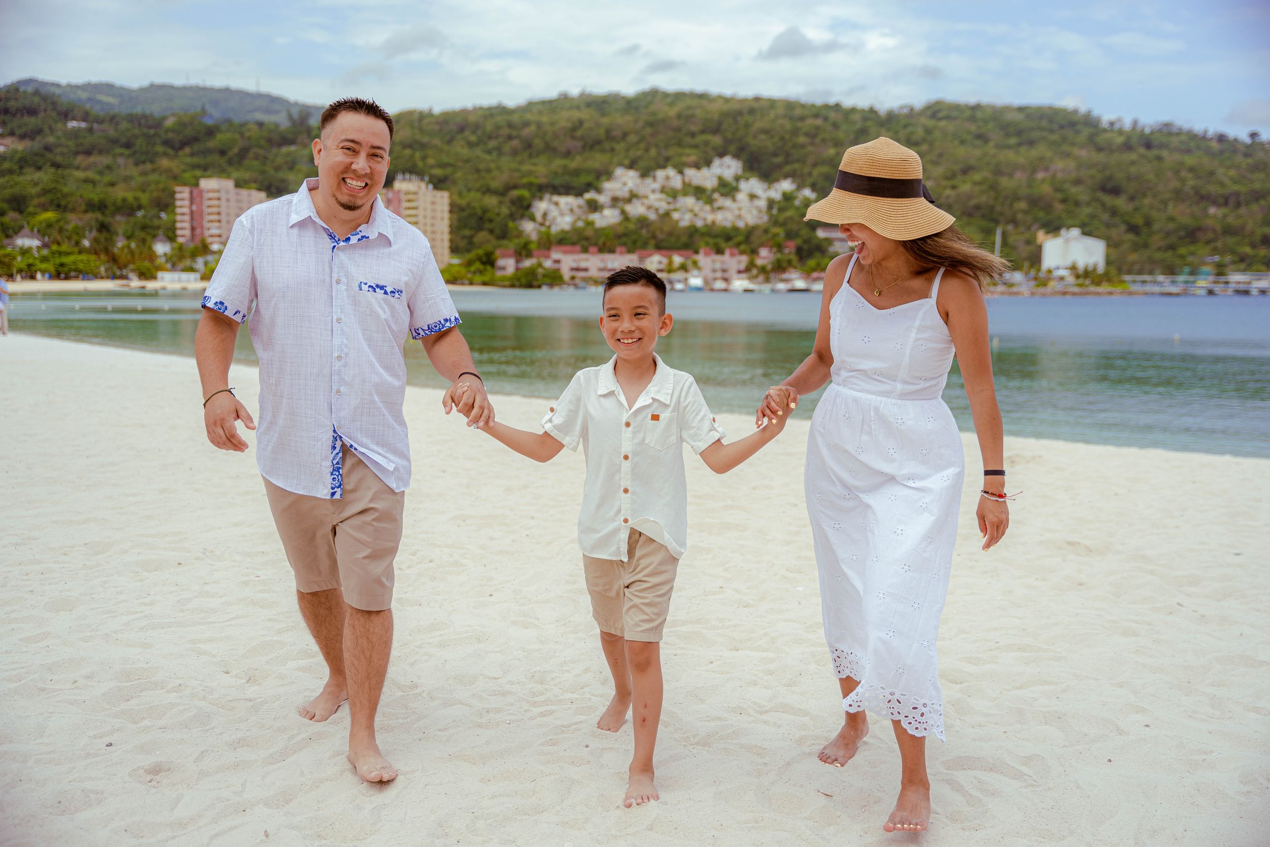 PJamaican Family Photographer Audley Green  eople walking together on a white sandy beach near calm waters and green hills on a sunny day.