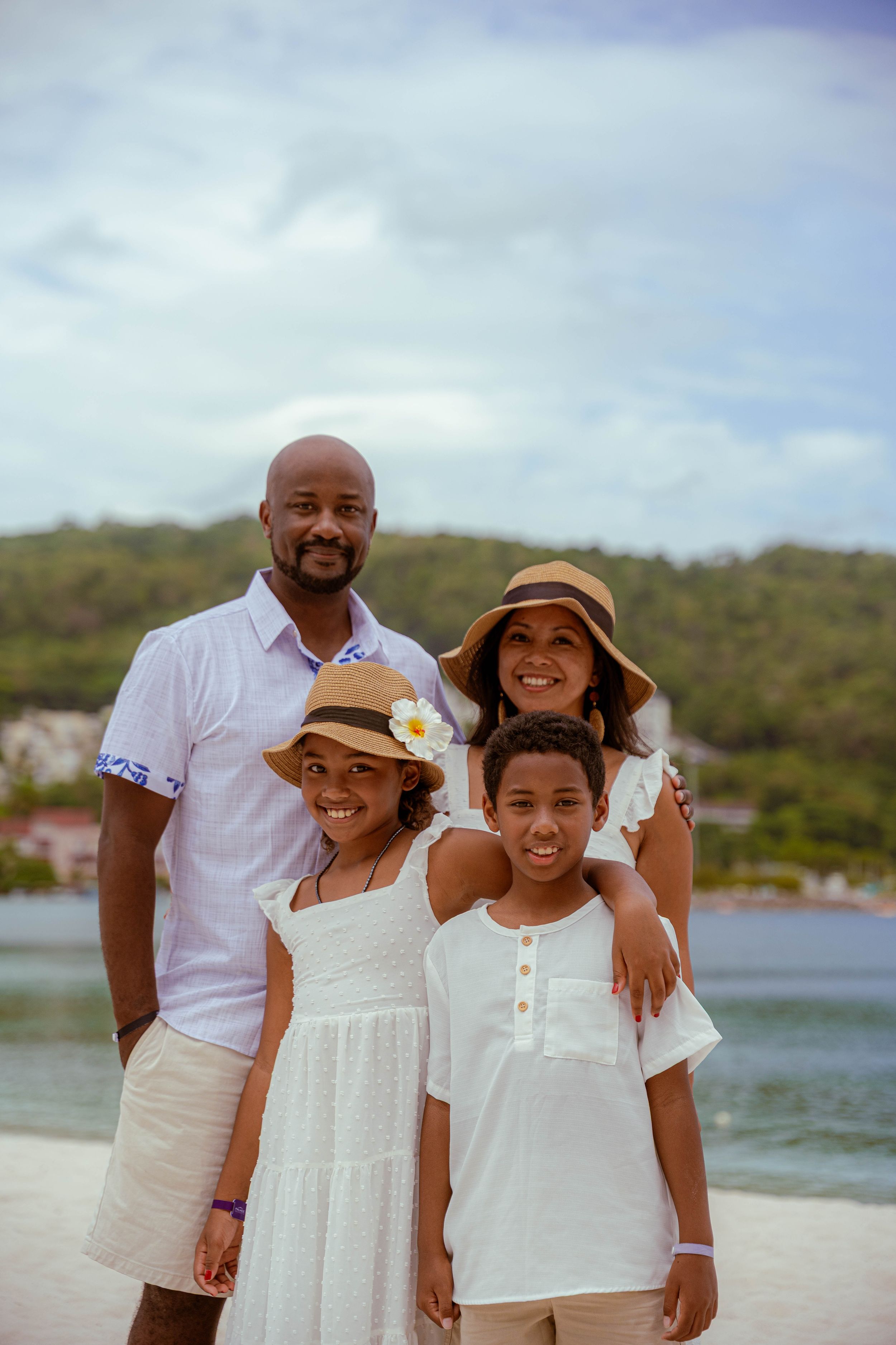 Jamaican Family Photographer Audley Green  Family of four in coordinated white outfits posing on beach with sun hats and mountain scenery.