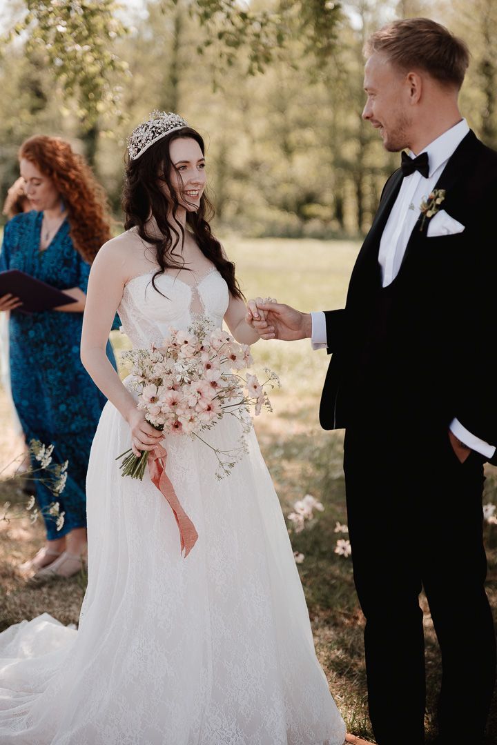 A bride in a white gown and tiara shares a joyful moment with a groom in black tuxedo during an outdoor wedding ceremony.