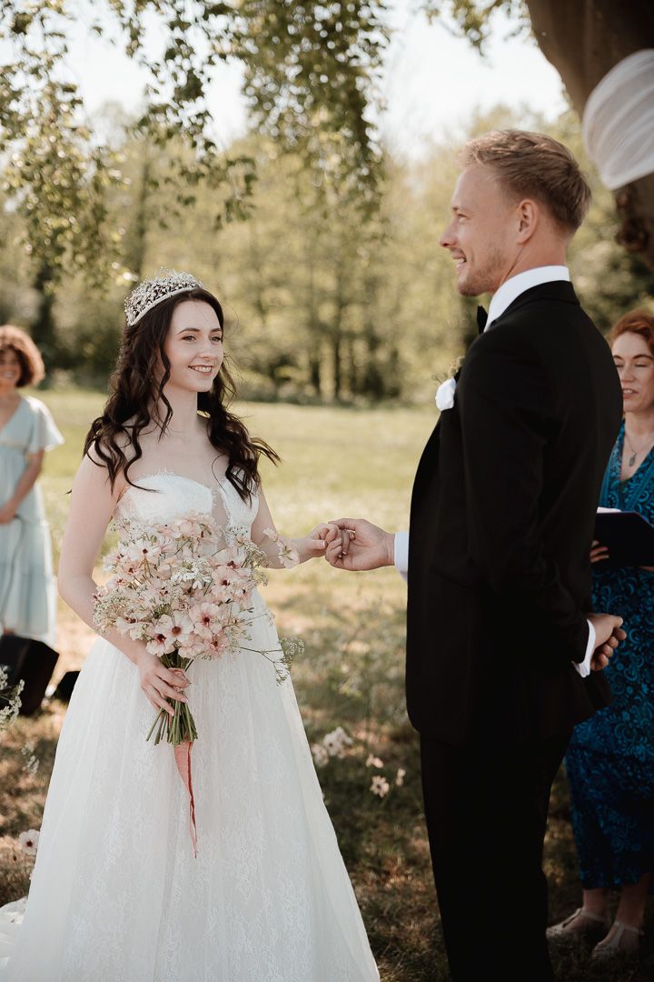A bride in a white gown and tiara holds a pink bouquet while exchanging vows during an outdoor ceremony.