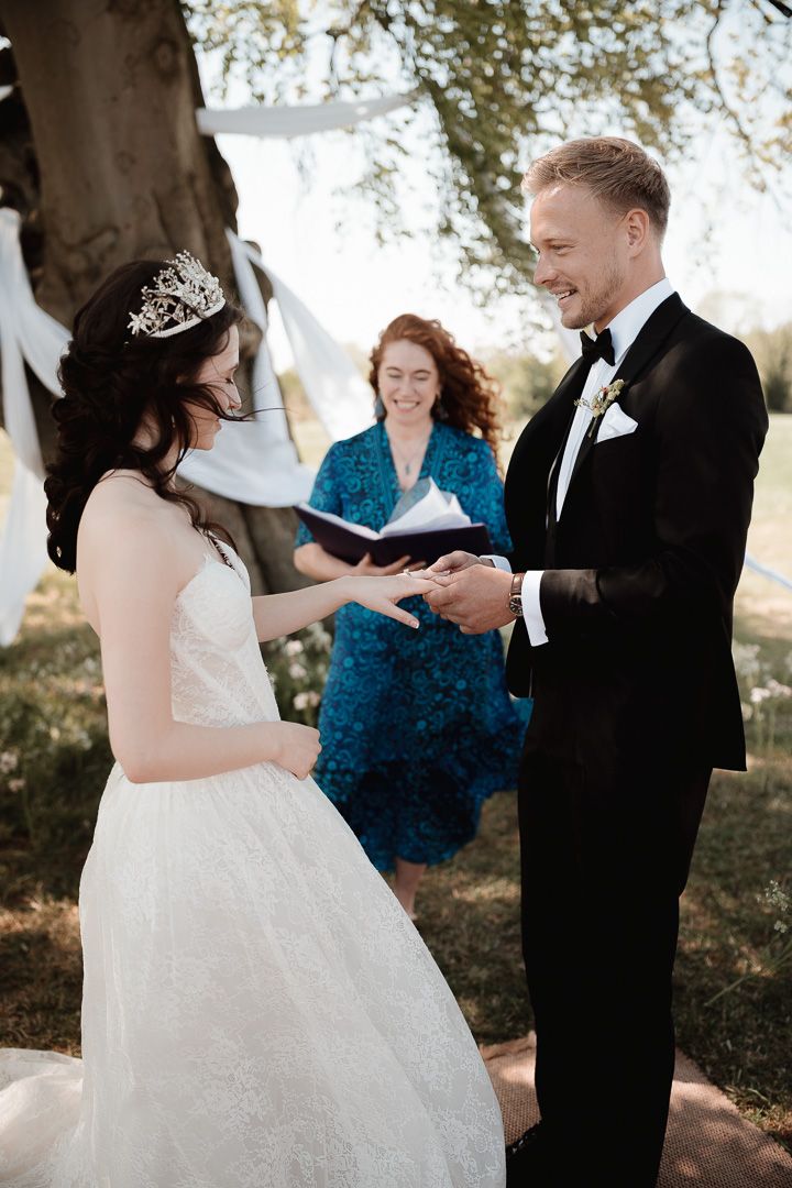 Romantic wedding couple exchanging rings during outdoor ceremony under sunlit trees.