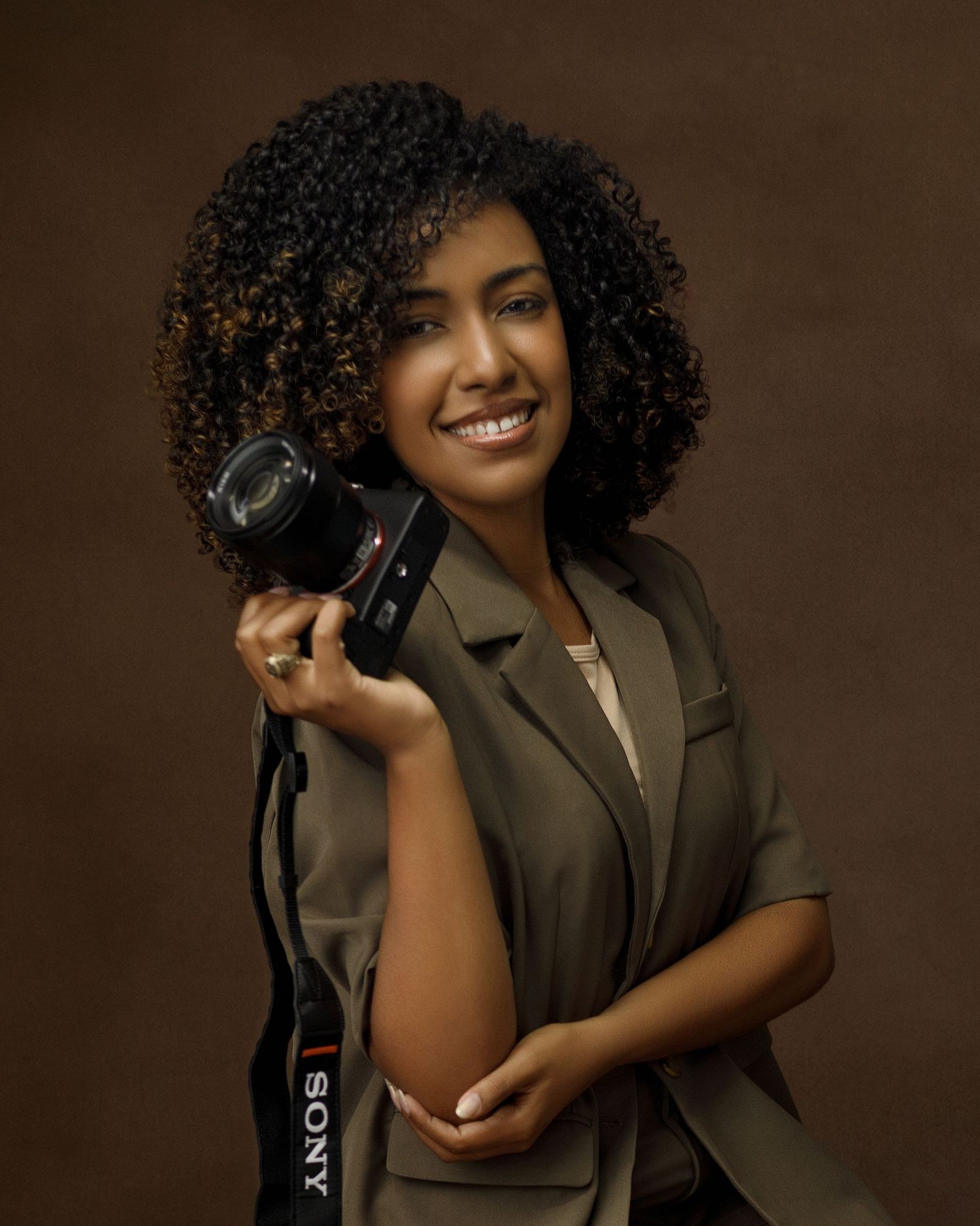 Professional photographer in olive blazer holding camera against brown backdrop.
