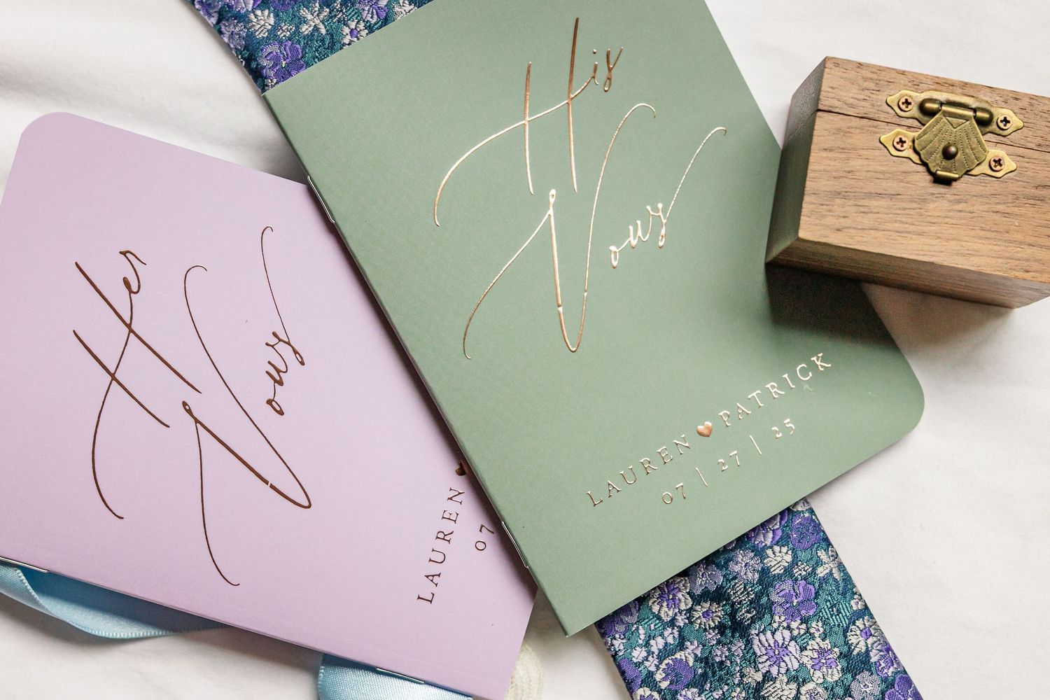 Wedding invitation cards in pastel green and pink with gold foil lettering laid on floral patterned paper background.