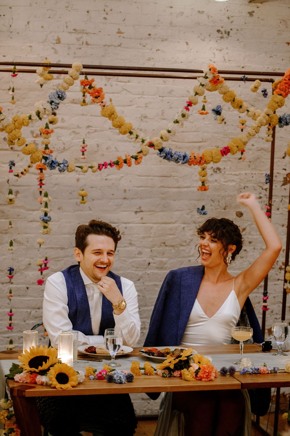 A joyful wedding reception moment captured at a rustic brick wall venue with colorful hanging decorations.