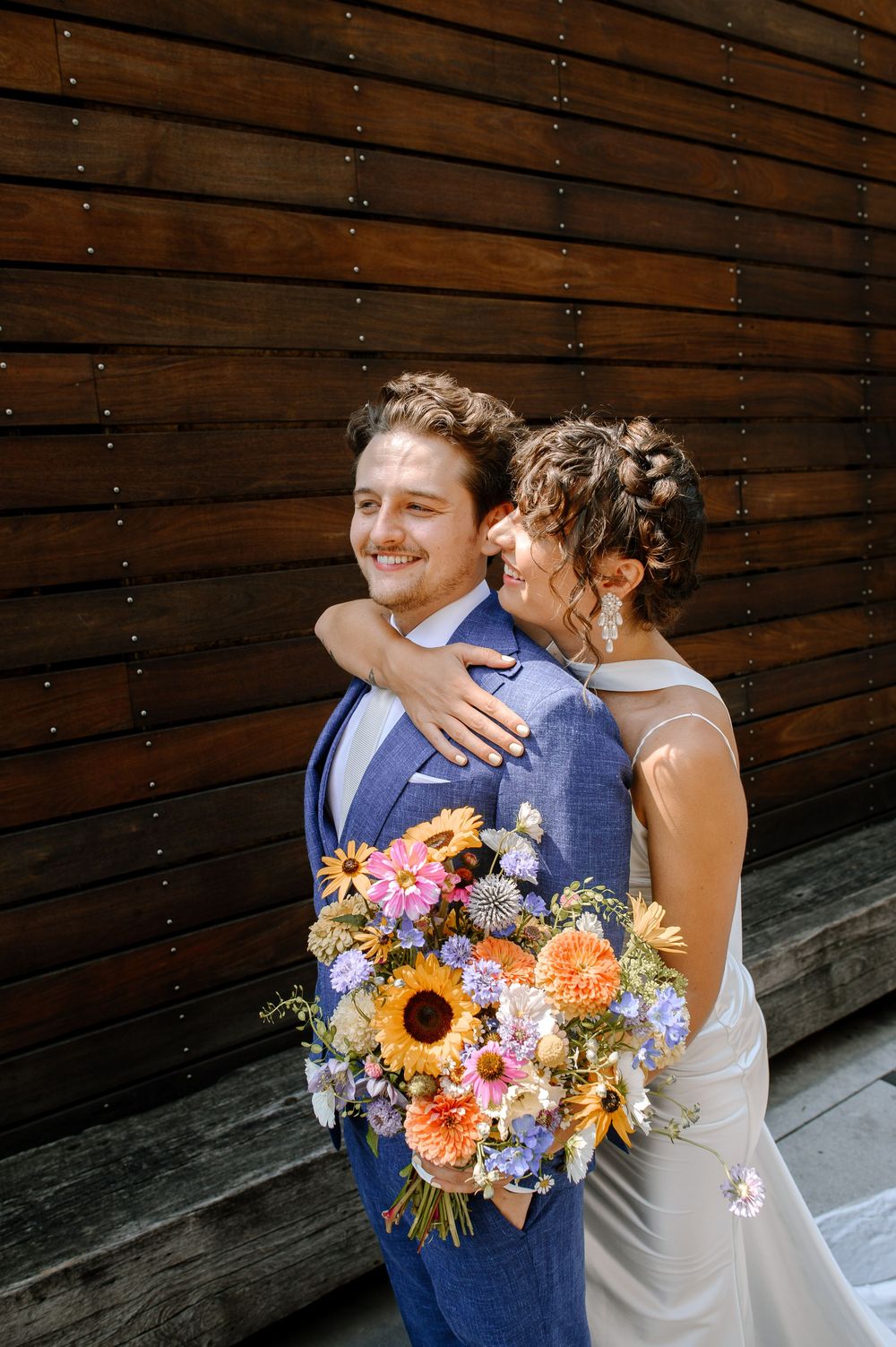 Couple embraces in front of wooden wall while holding vibrant bouquet with sunflowers and pink blooms on sunny day.