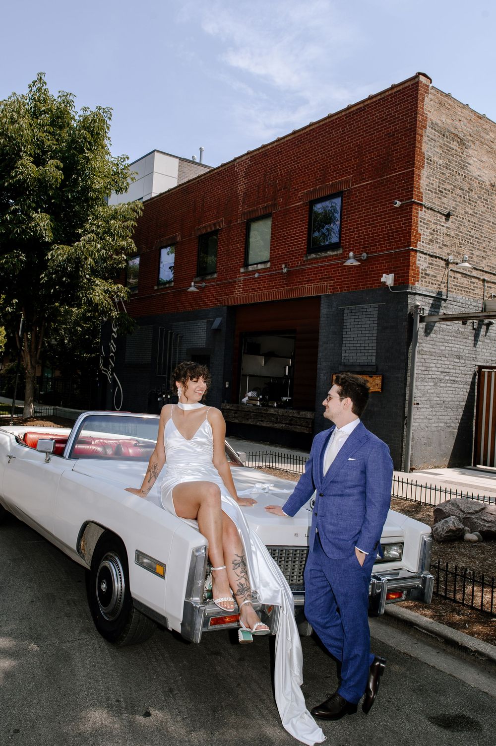 Newlyweds share romantic moment beside classic white vintage car on urban street.