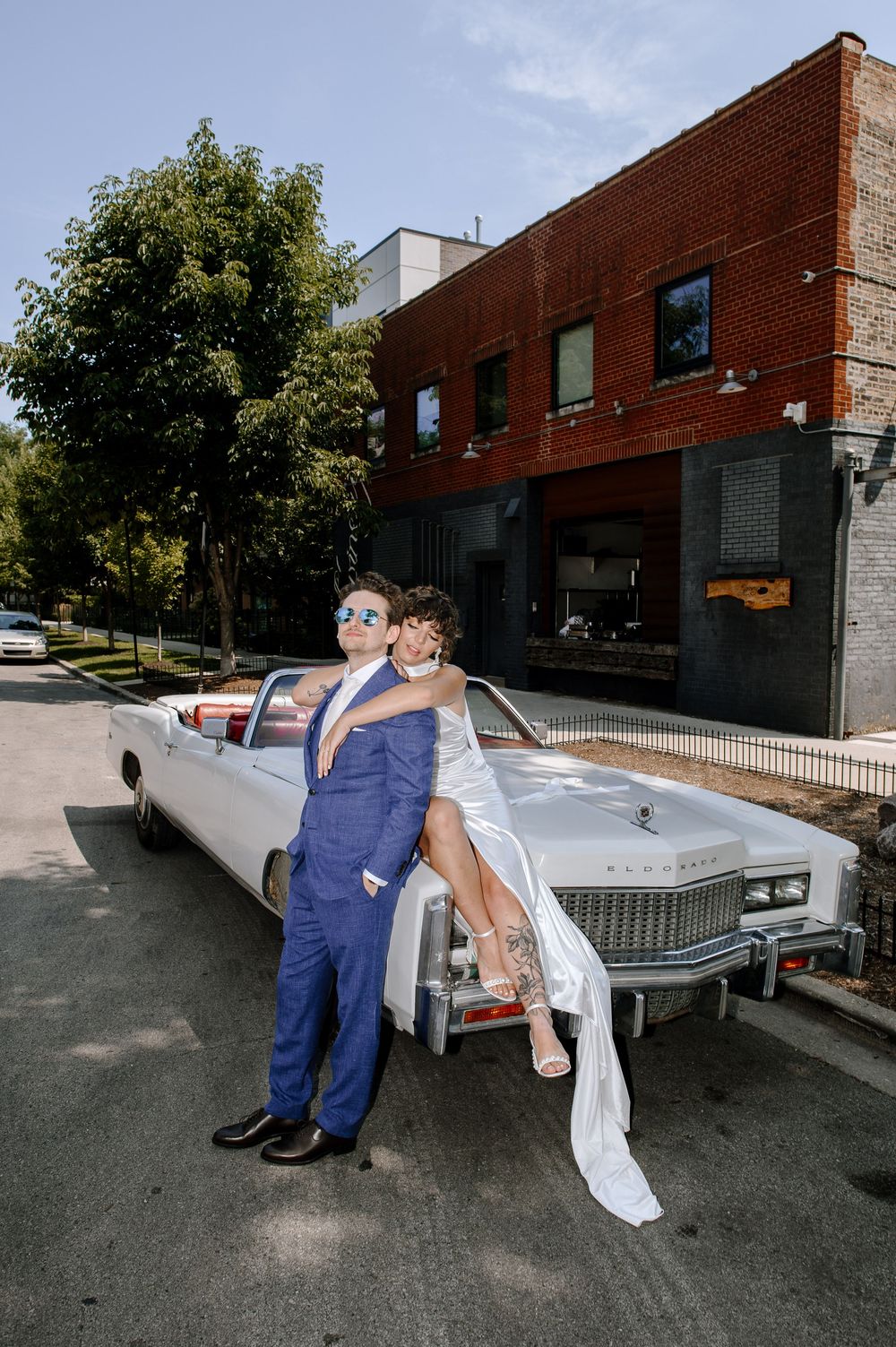 A classic white Cadillac parked in front of a brick building serves as backdrop for a stylish wedding photo.