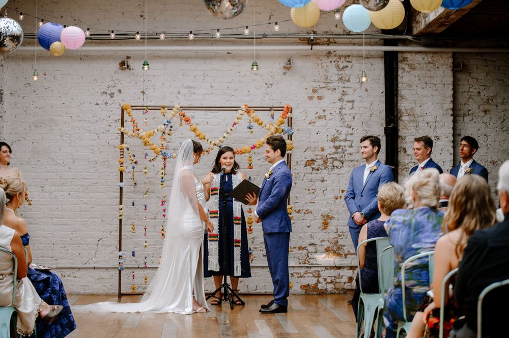 Indoor wedding ceremony takes place in an industrial-style venue with exposed brick walls and hanging decorations.