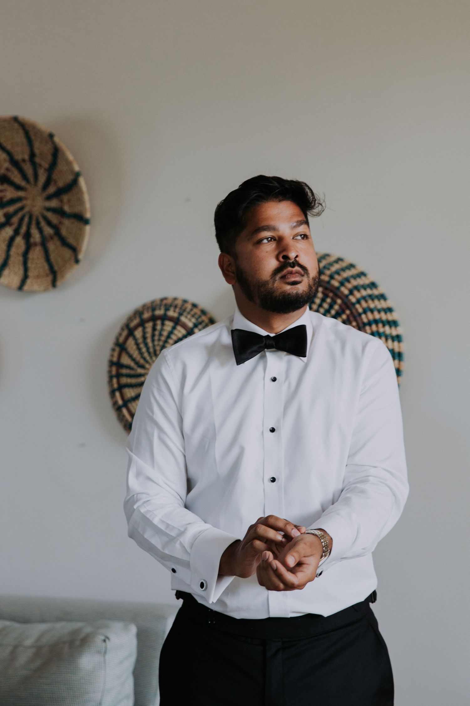 Groom in classic tuxedo and bow tie stands against wall decorated with woven art pieces.