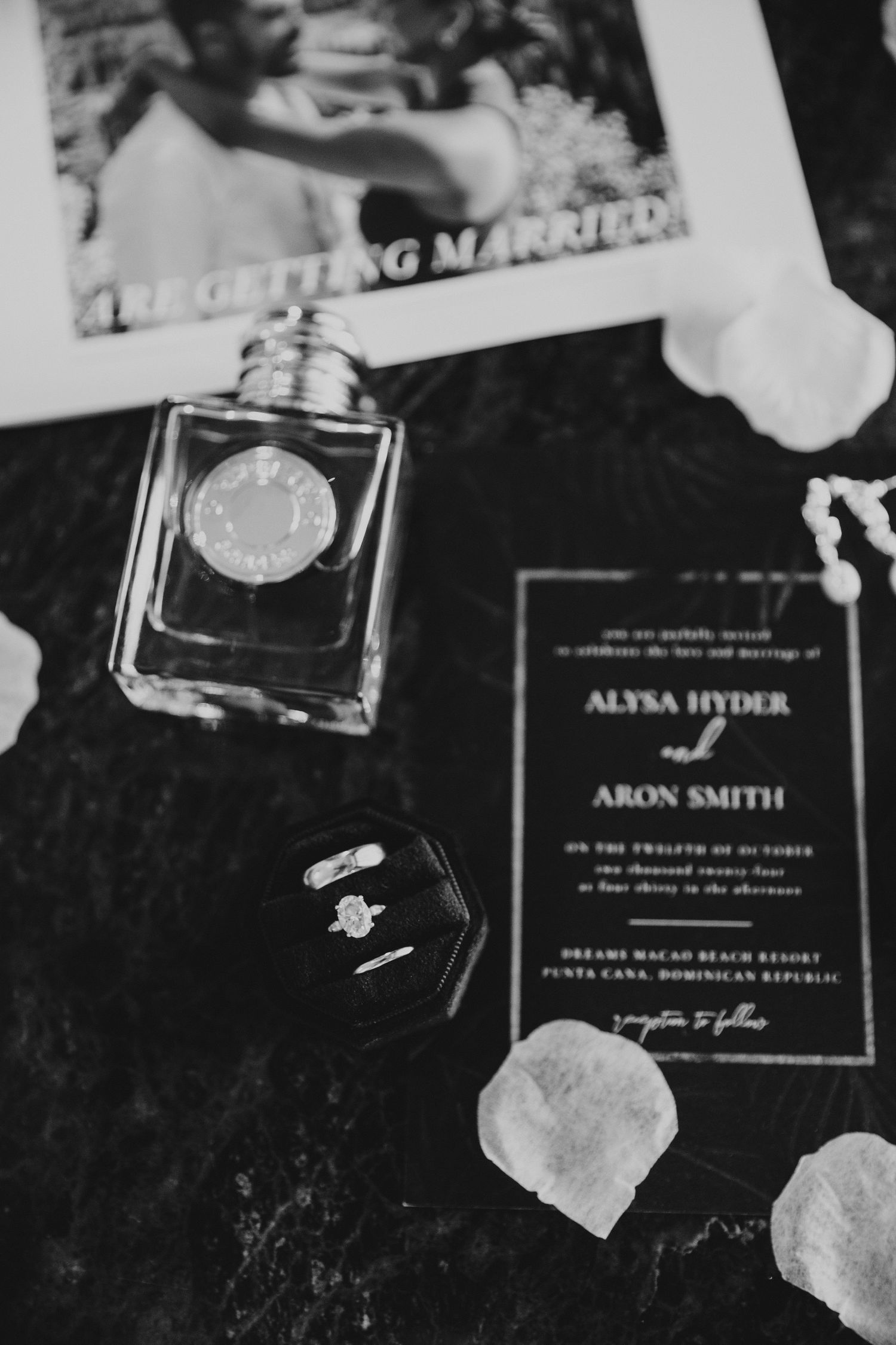 Wedding invitation and perfume bottle arranged on dark surface with scattered petals in black and white.