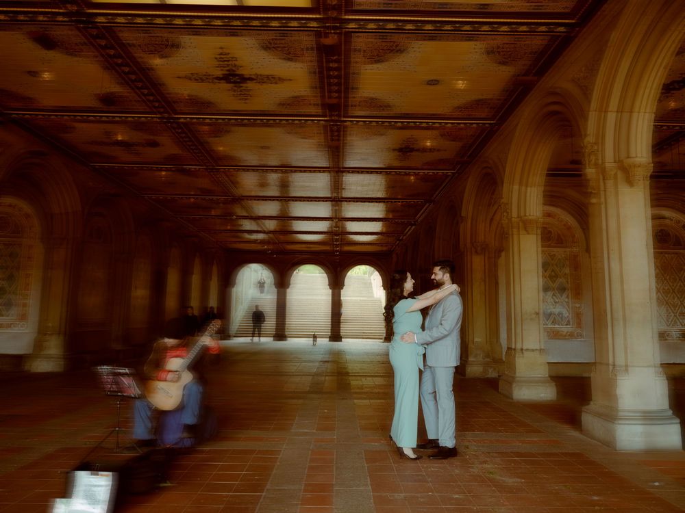 A romantic couple embraces at Bethesda Terrace arcade in Central Park while a figure runs past them in the distance.