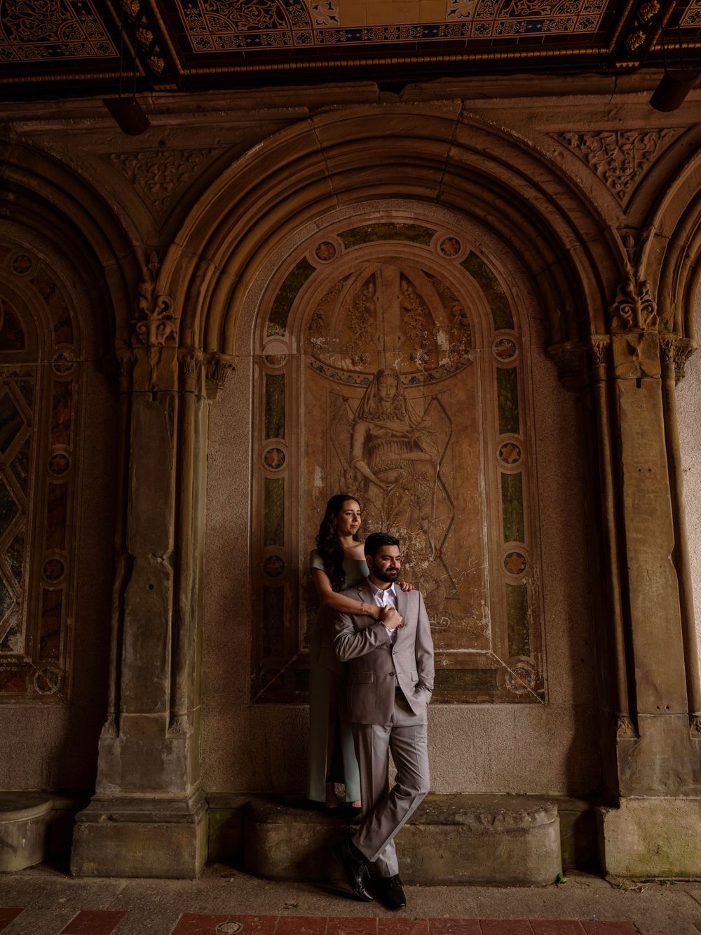 A couple poses intimately against ornate stone archways at Bethesda Terrace in Central Park with moody lighting.