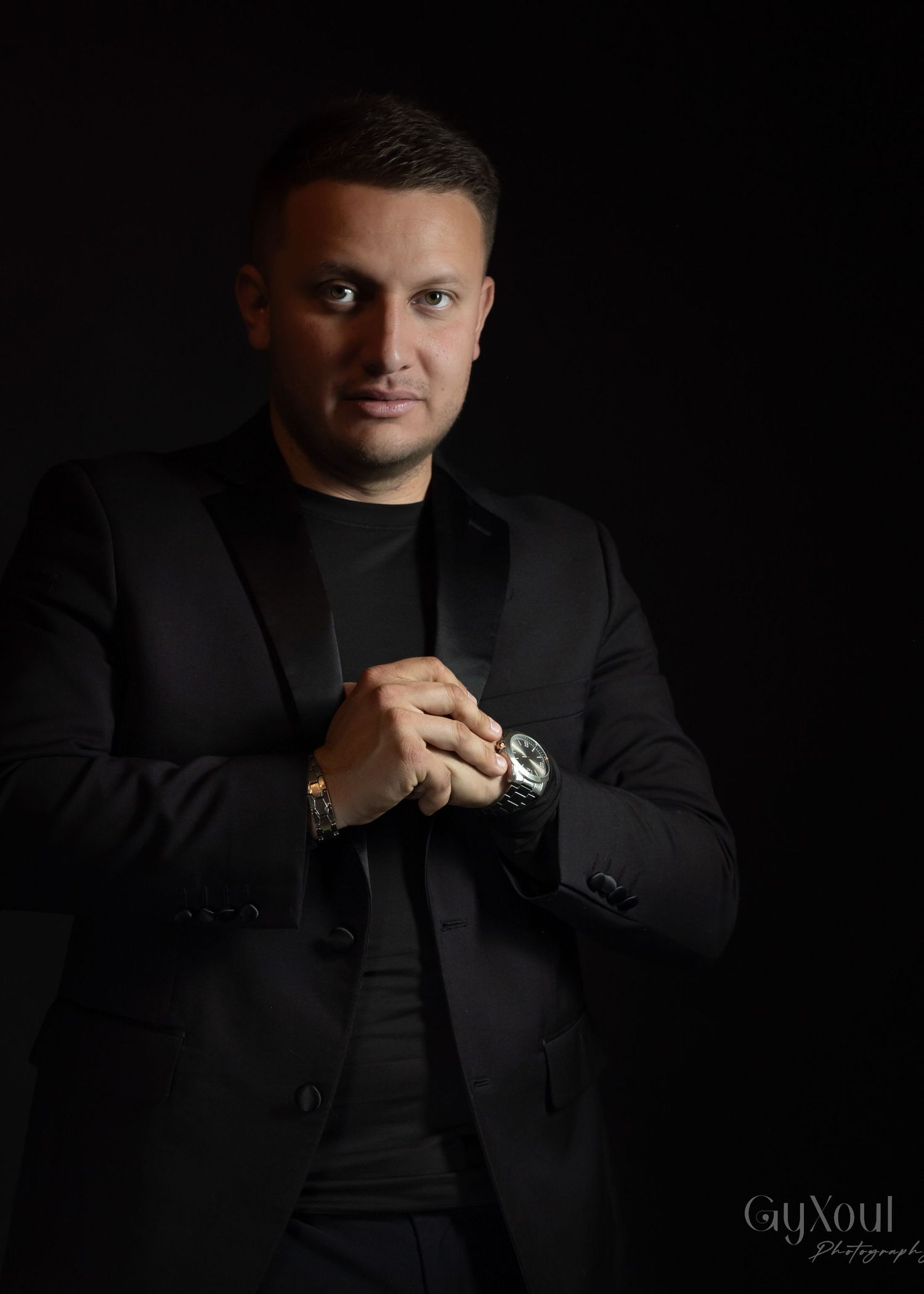 Professional studio portrait against dark background with subject wearing black suit and watch, styled in dramatic lighting.