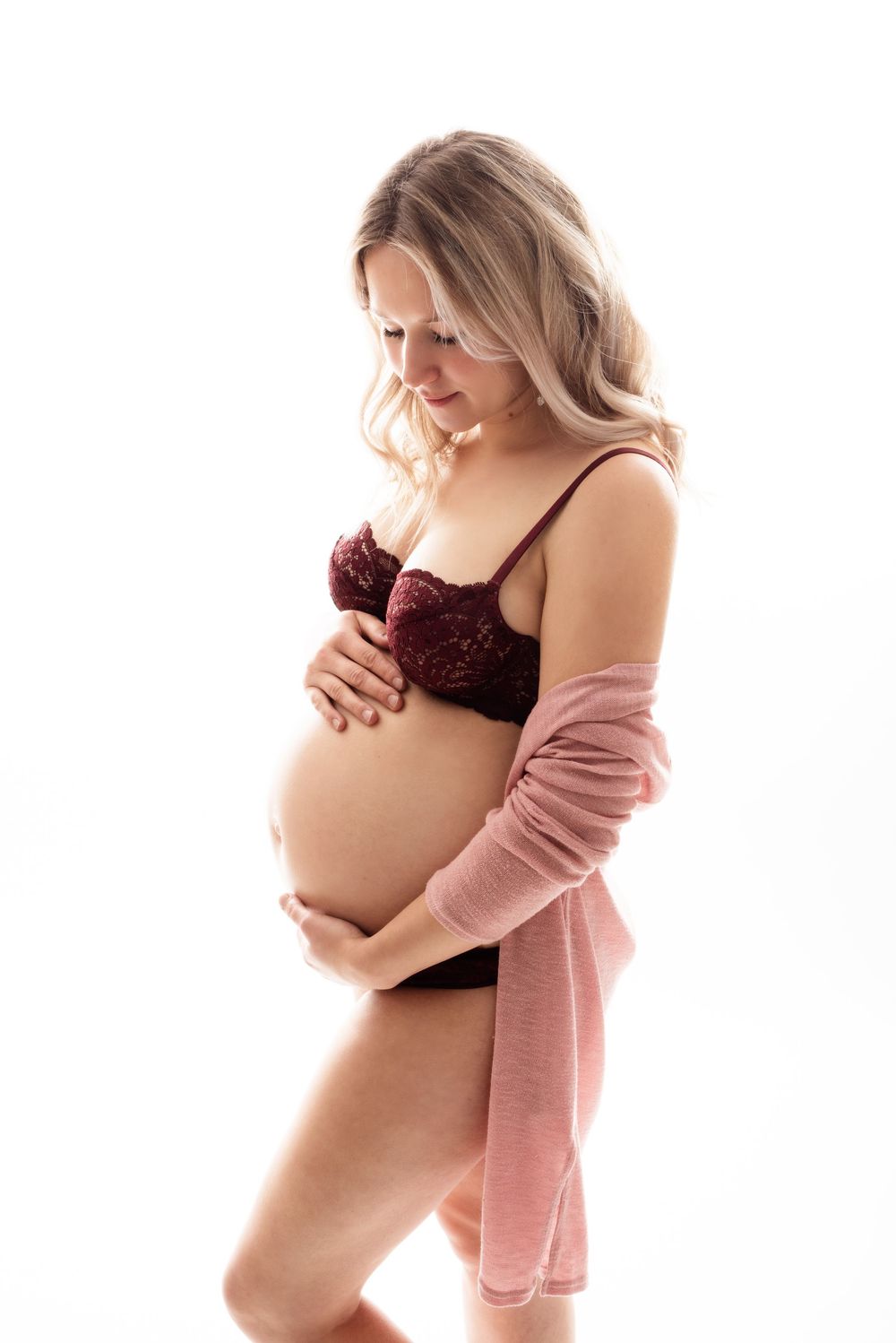 Maternity photo of expectant mother in burgundy lingerie and pink cardigan against white background.