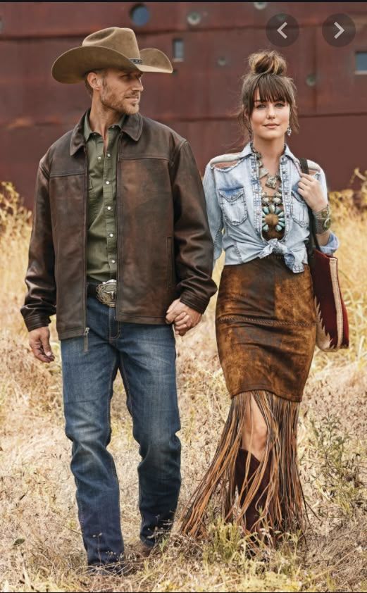 Western-styled couple walking together in dry grass field wearing cowboy hat, leather jacket and fringed suede skirt.