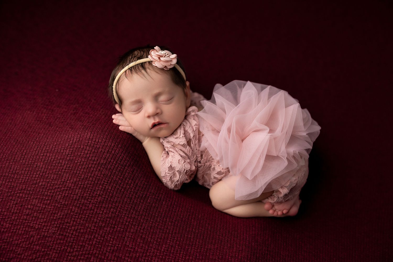 Sleeping newborn baby wrapped in dusty rose tulle, wearing floral headband, lying on pink textured backdrop.