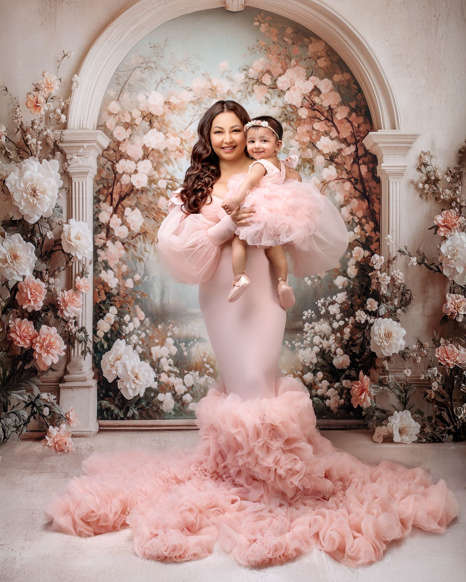 Mummy and me portrait of a woman embracing her daughter both smiling in matching designer peach gowns against a stunning paste arch backdrop