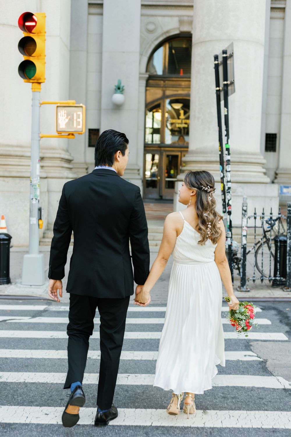 A couple in formal attire holding hands while walking across a city crosswalk at a stoplight.