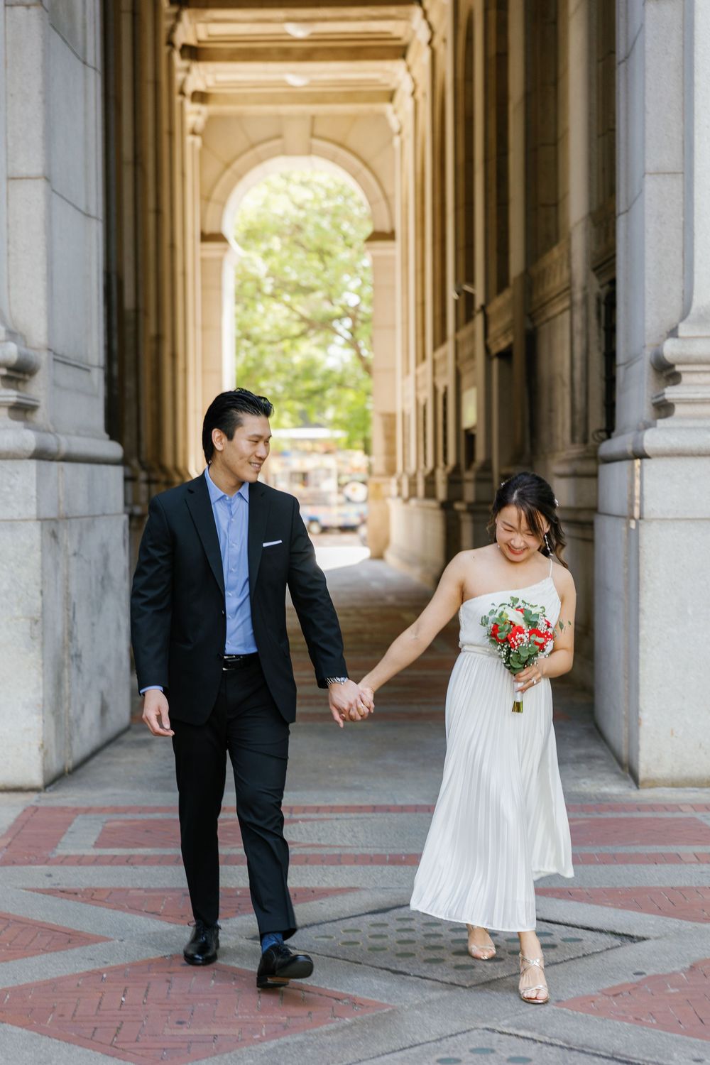 A couple in wedding attire walks through an elegant stone archway colonnade holding hands on a sunny day.