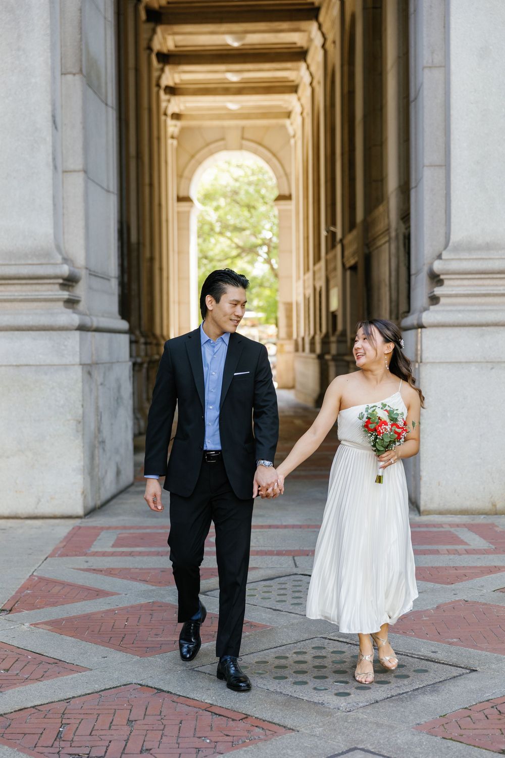 Couple holds hands while walking through an elegant stone archway in a romantic wedding photoshoot.