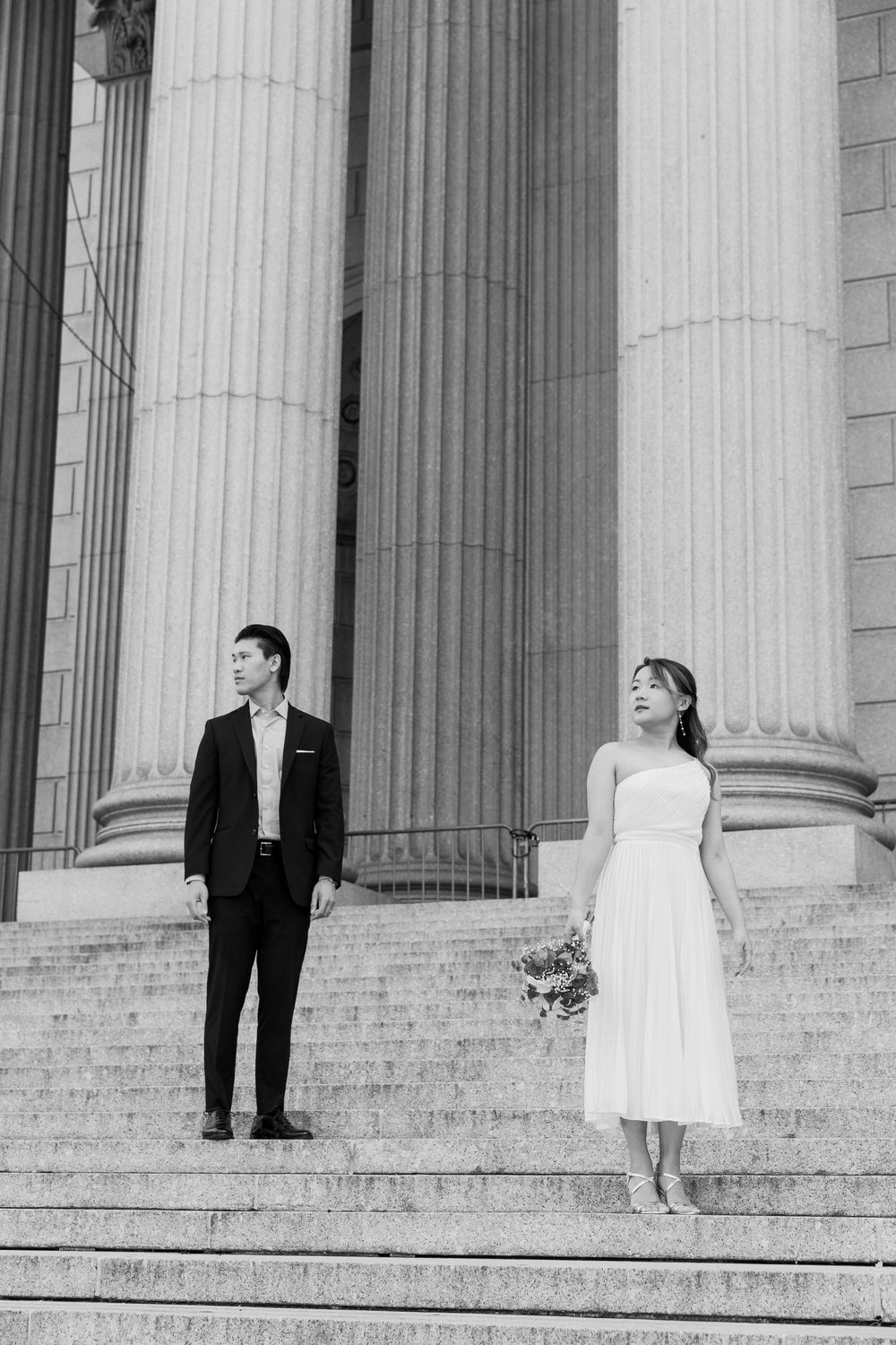 A couple poses on grand marble steps with white columns for wedding photos in black and white.