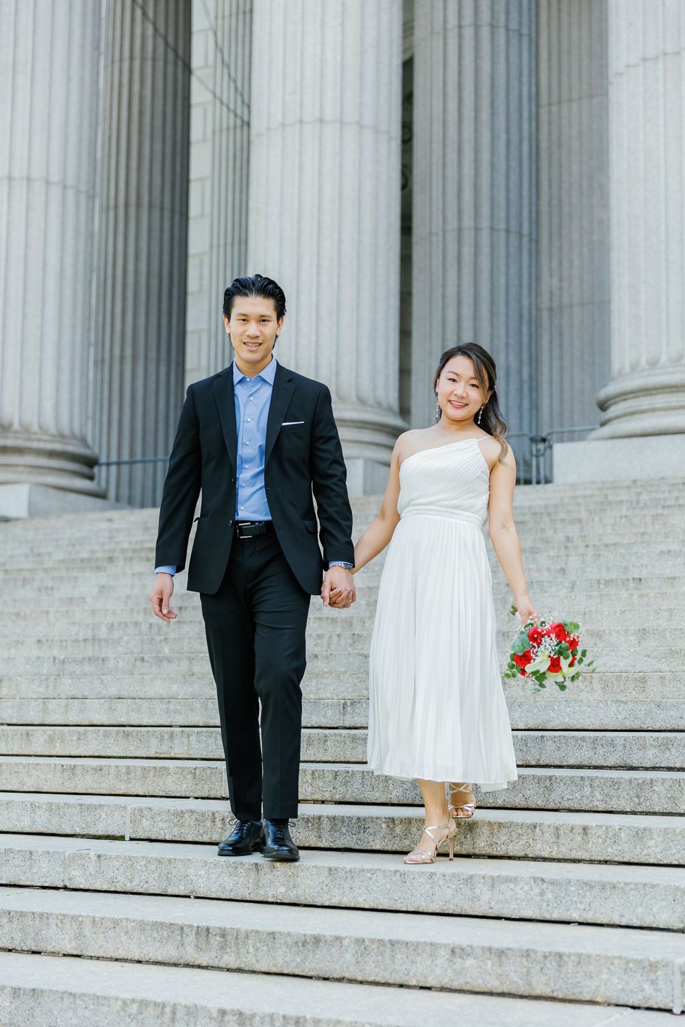 A couple descends the grand marble steps of a classic architectural building with columns, dressed in formal wedding attire.
