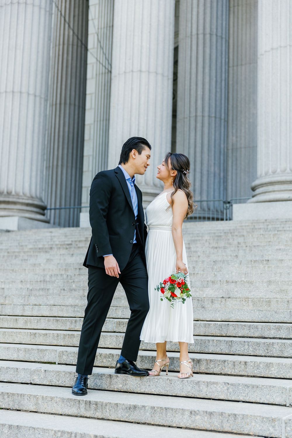 A romantic wedding couple poses on grand marble steps with classical columns in the background.