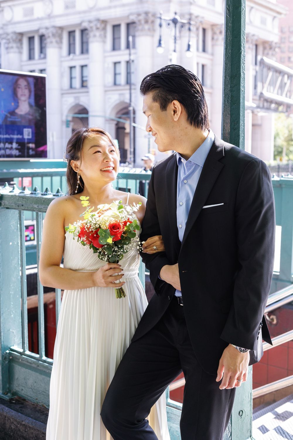 A couple in wedding attire share joyful moments together on a balcony with white railing and buildings in the background.