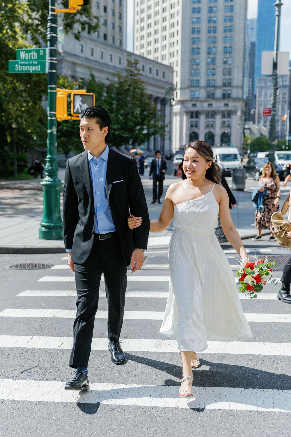 A newlywed couple walks across a city crosswalk in Manhattan with a bouquet of flowers.