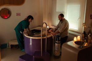 Medical professionals in green scrubs attend to a patient in a dimly lit room with purple birthing tub.