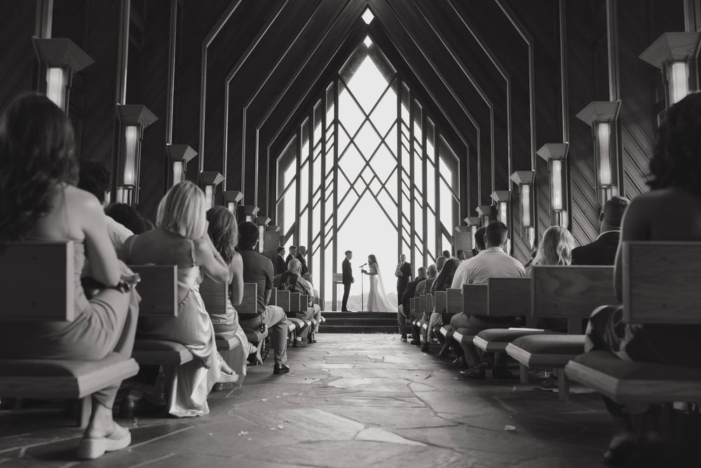 Black and white interior view of a chapel wedding ceremony with guests seated in pews and couple silhouetted at the altar.