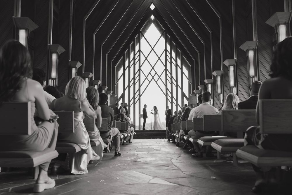 Black and white photograph of a wedding ceremony inside a chapel with dramatic lighting through gothic windows.