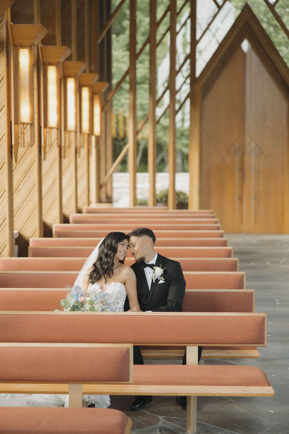 A romantic moment captured between couple sitting on wooden church pews in a beautiful sunlit chapel setting.