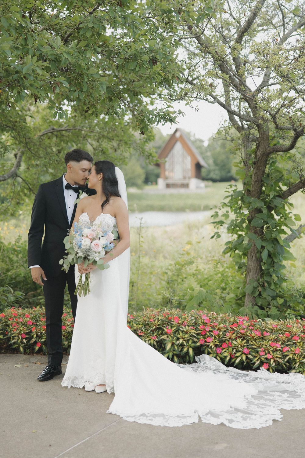 A couple poses in an outdoor garden setting with blooming red flowers and lush greenery.