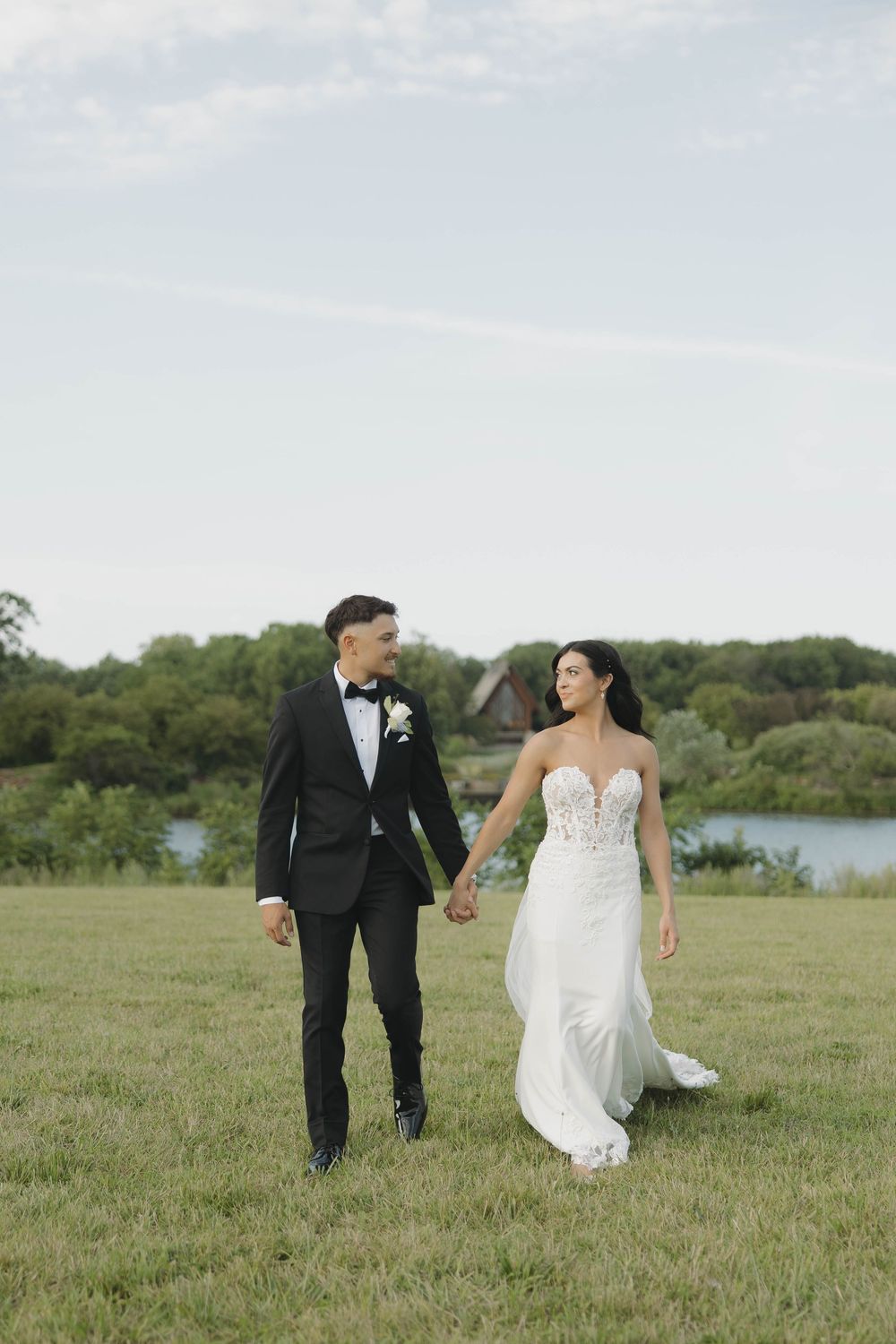 Wedding couple walking together in a scenic meadow with a lake and trees in the background.
