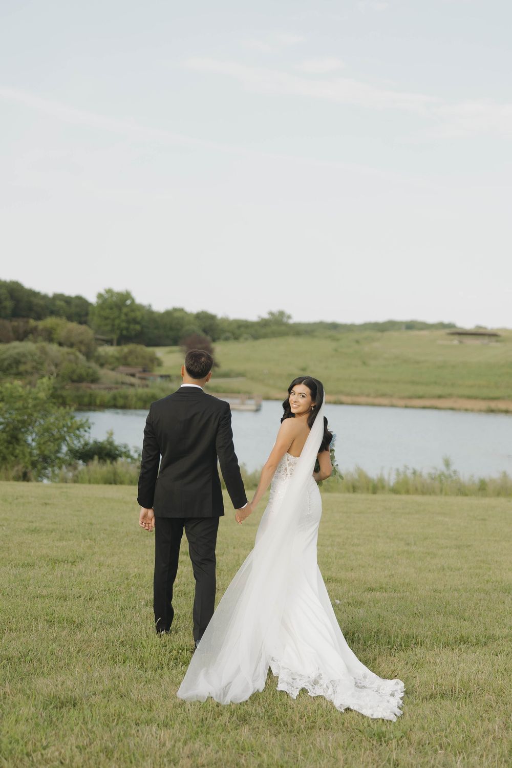 A couple walks through a serene meadow with a lake in the background on their wedding day.