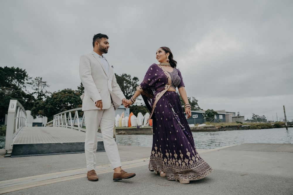 A romantic couple in traditional Indian wedding attire poses elegantly on a waterfront walkway under cloudy skies.