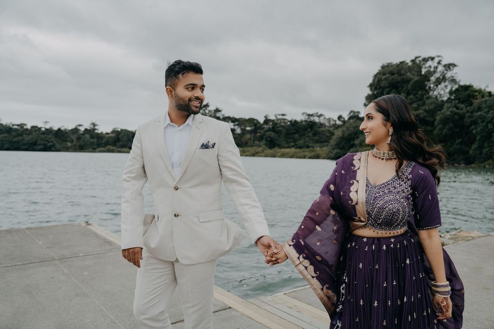 A couple holding hands while walking along a waterfront, with the person on the left in white and right in purple attire.
