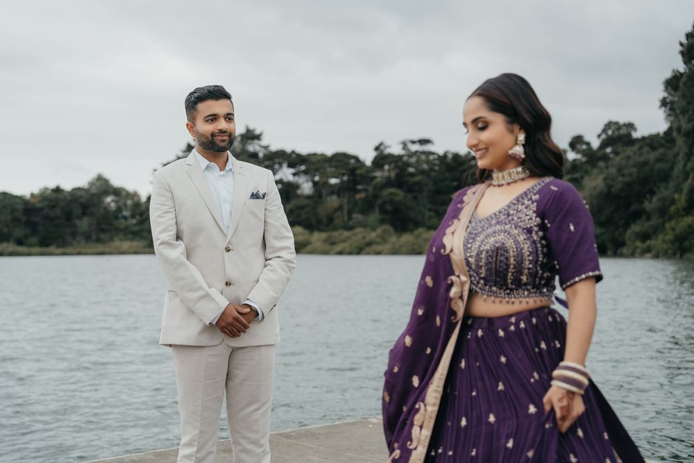 A couple in formal attire stands by a lake, with one person wearing a beige suit and another in a purple traditional dress.