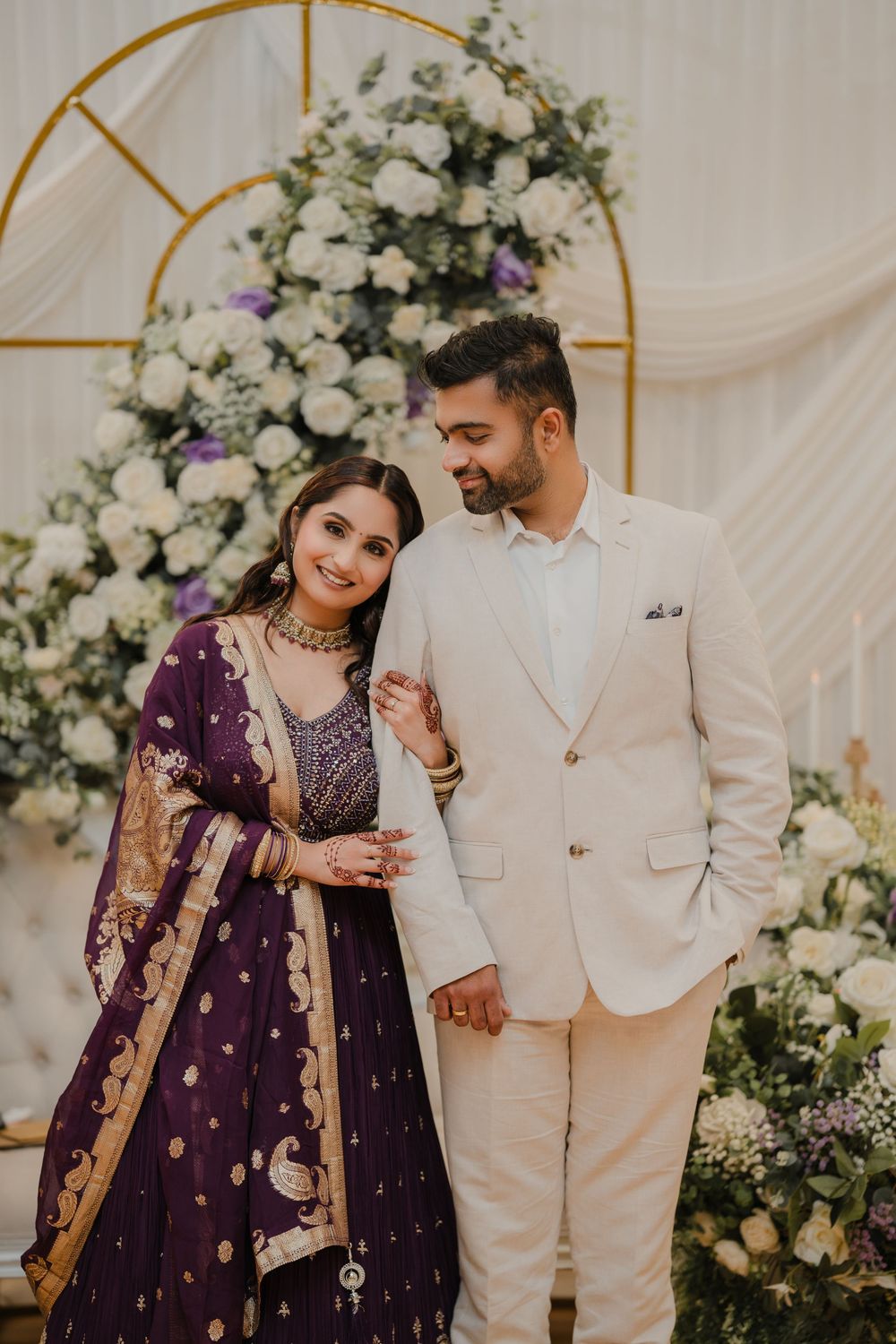 Elegant couple in traditional Pakistani wedding attire share romantic moments against floral backdrop in white and purple decor.