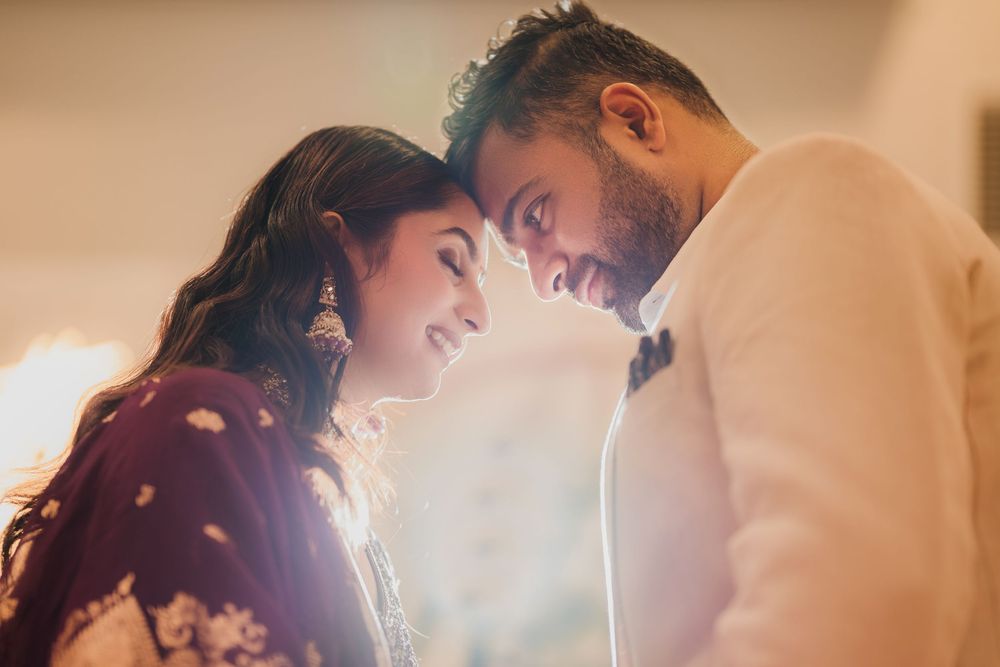 A romantic couple in traditional Indian wedding attire share an intimate moment with foreheads touching in warm lighting.