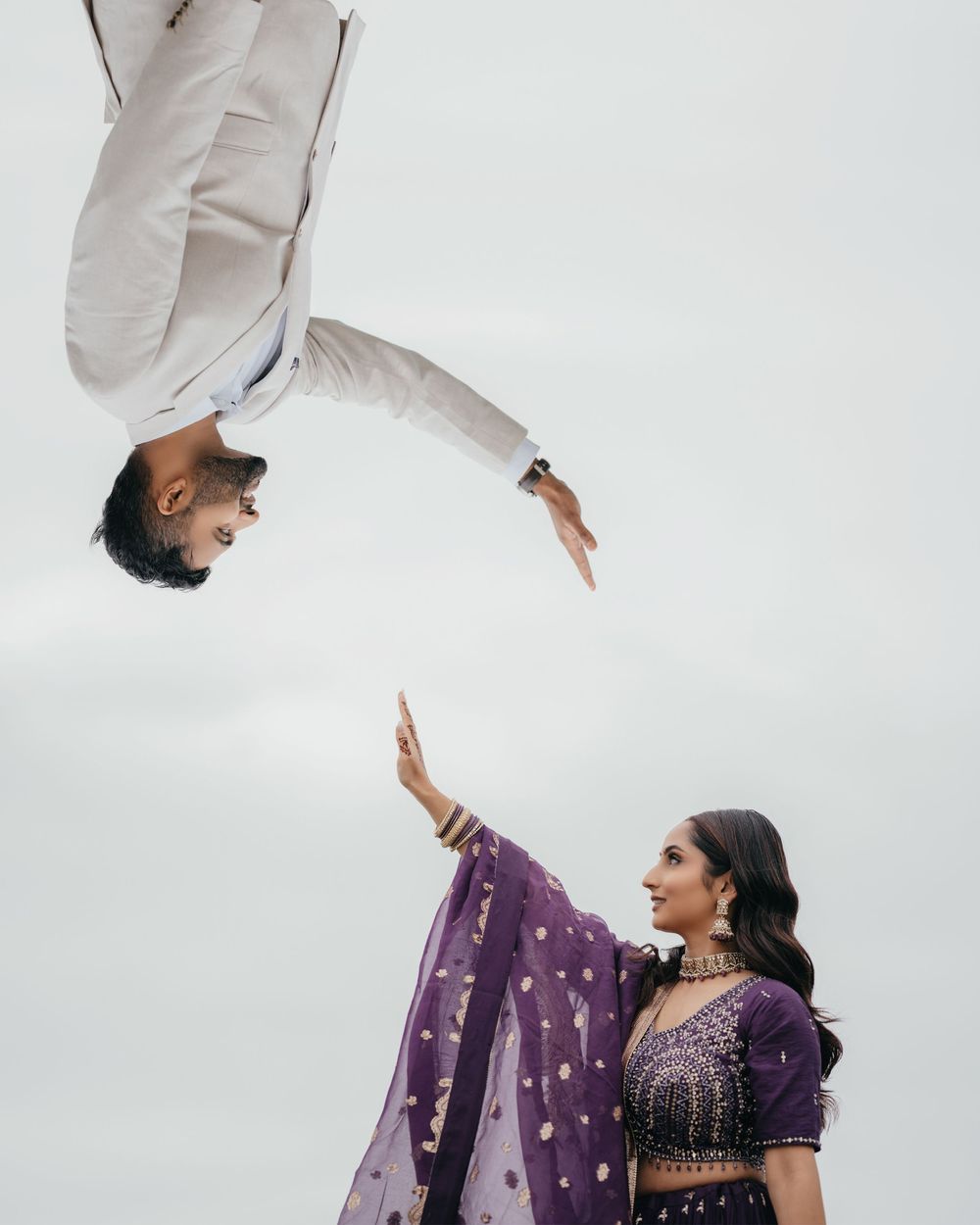 Creative wedding photo with groom floating above bride in purple lehenga against white background.