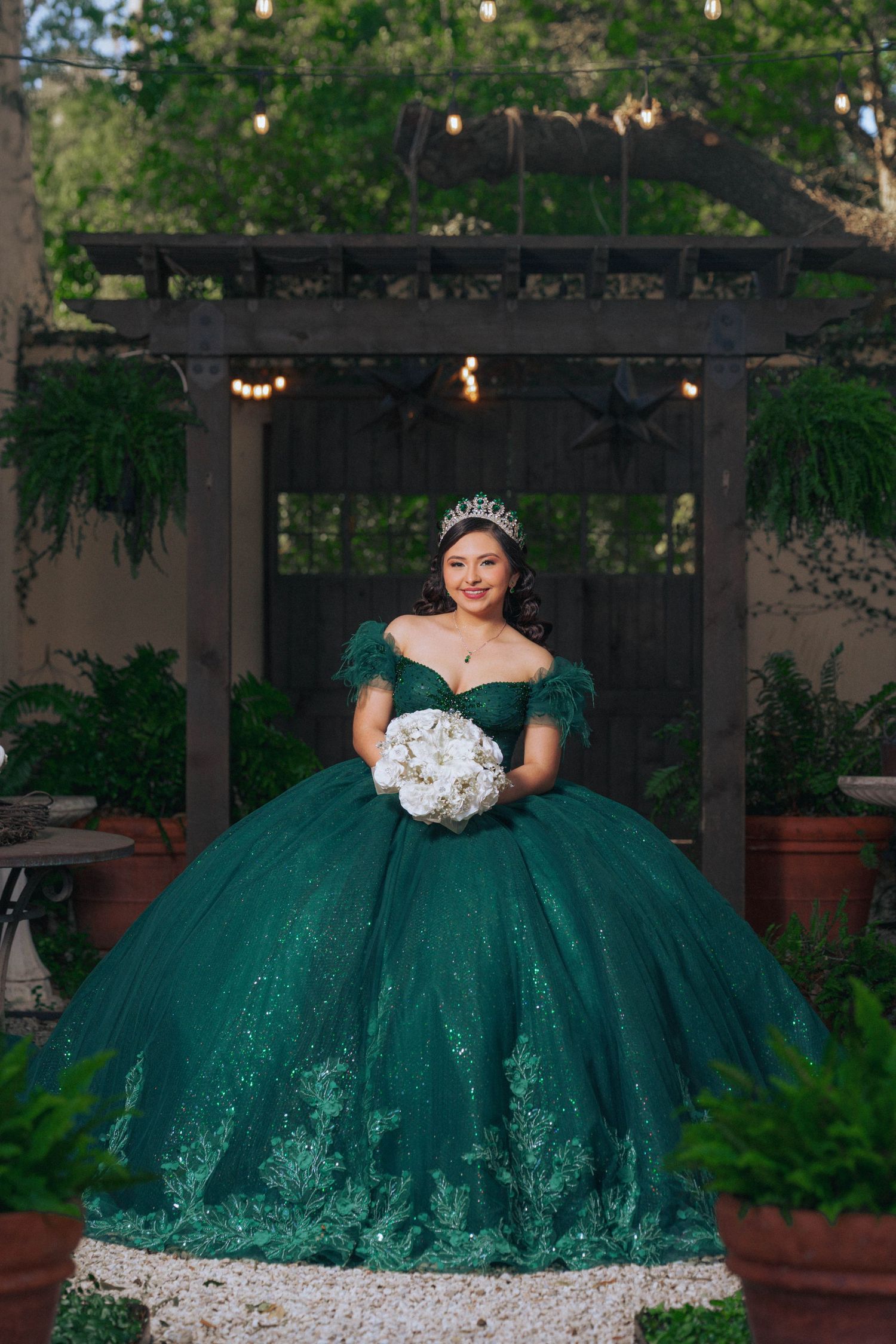 A stunning emerald green quinceañera ball gown photographed in a garden setting with string lights.