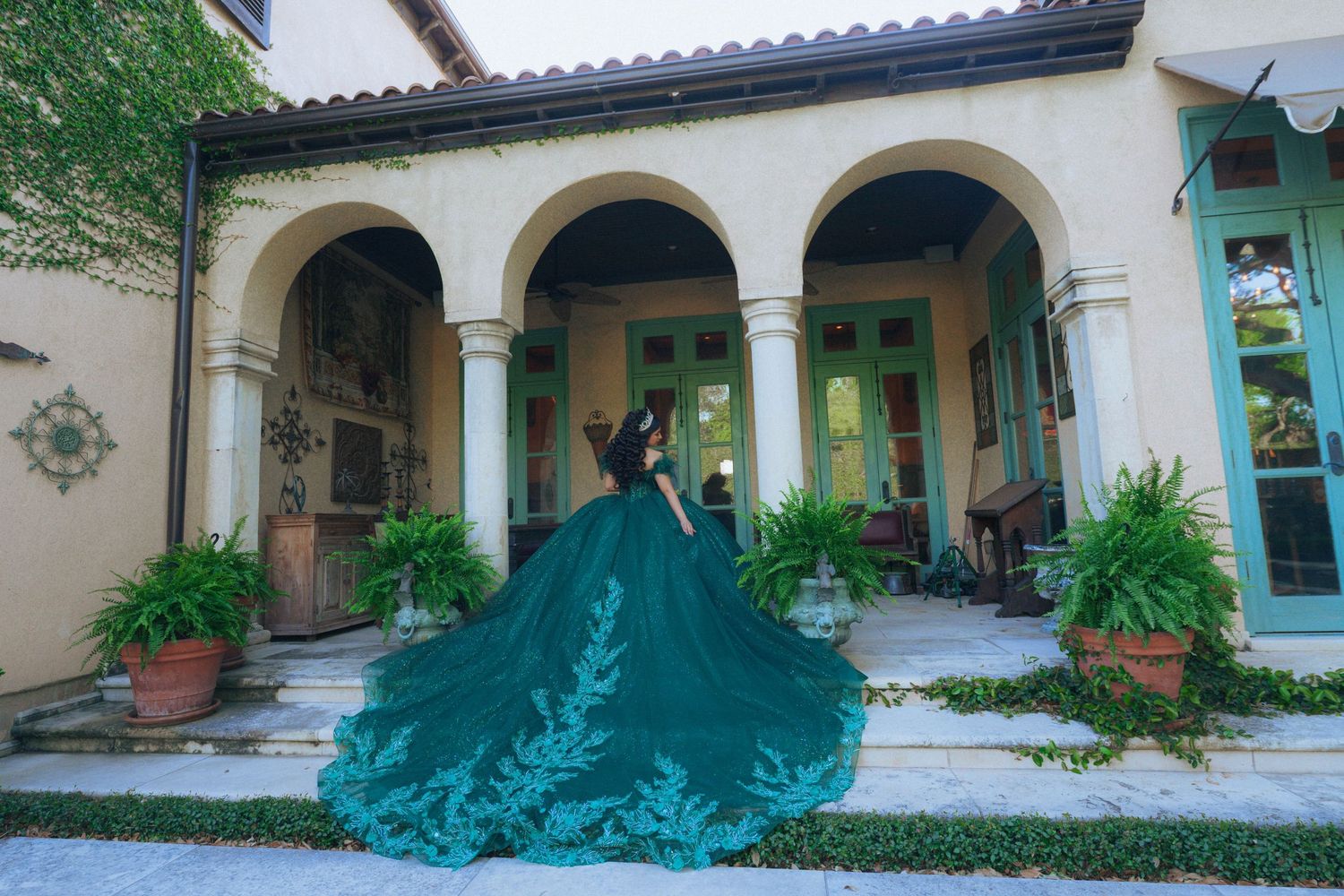 Elegant emerald gown flowing across Spanish-style courtyard with white archways and terracotta tiles.