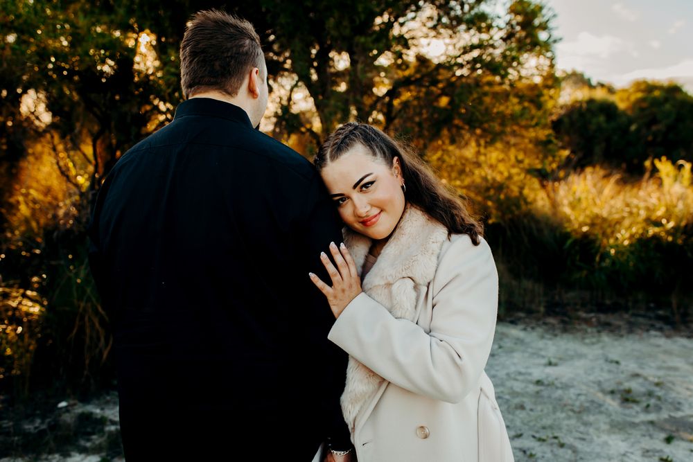 A couple in wedding attire embraces during golden hour sunset at a beachside location.
