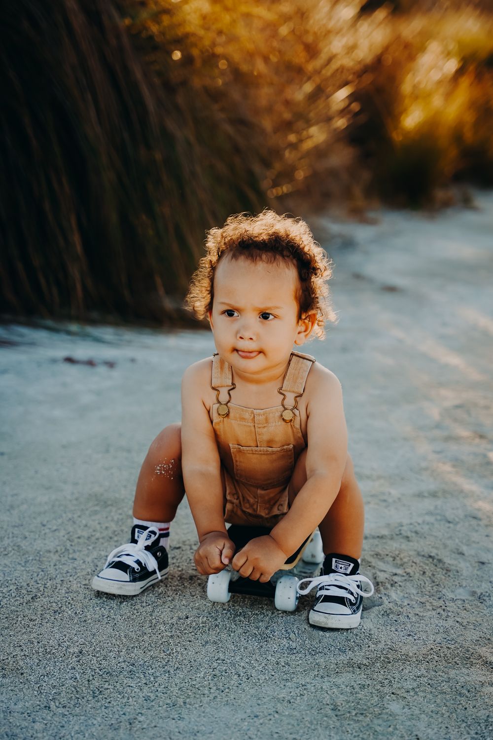 Small child wearing brown overalls and black sneakers sits and crawls on sandy ground during golden hour.