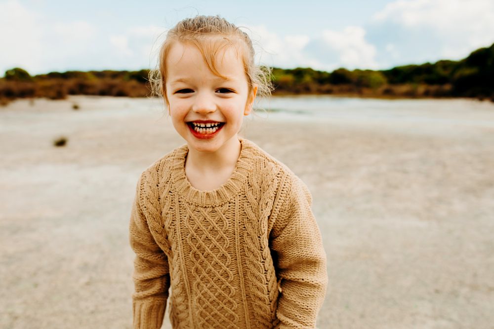 A laughing child wearing a cozy tan cable knit sweater stands on a sandy beach at golden hour.